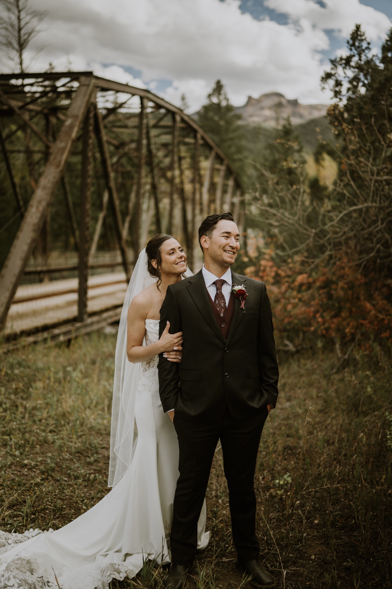 Bride and groom standing near a rustic bridge with mountains in the background